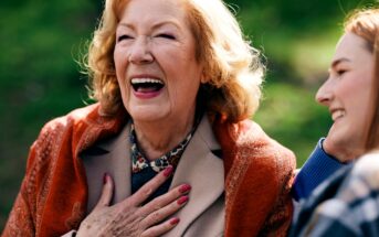 An elderly woman with blonde hair and a bright smile is laughing heartily with her hand on her chest. She is wearing a beige coat with an orange scarf. A younger woman next to her is smiling, looking at her. They are outdoors with a blurred green background.