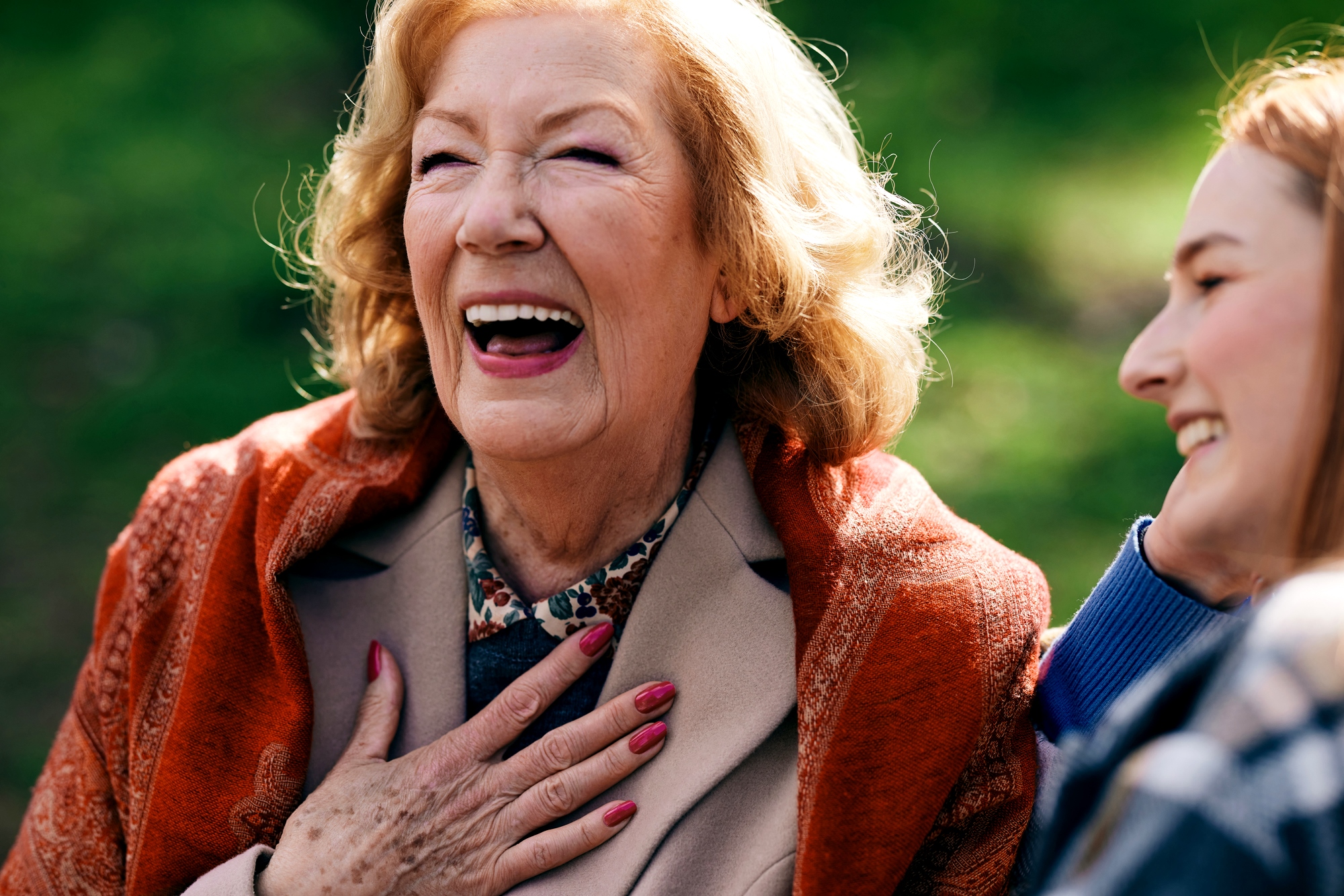 An elderly woman with blonde hair and a bright smile is laughing heartily with her hand on her chest. She is wearing a beige coat with an orange scarf. A younger woman next to her is smiling, looking at her. They are outdoors with a blurred green background.