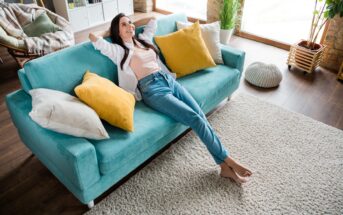 A woman is lying on a turquoise couch with her arms stretched behind her head, smiling. The living room features large windows, a light-colored rug, and several pillows in shades of white and yellow. Wooden flooring and indoor plants complement the cozy atmosphere.