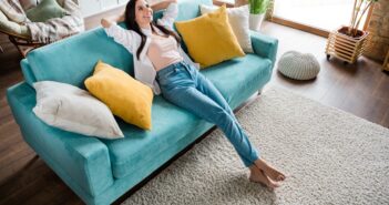 A woman is lying on a turquoise couch with her arms stretched behind her head, smiling. The living room features large windows, a light-colored rug, and several pillows in shades of white and yellow. Wooden flooring and indoor plants complement the cozy atmosphere.