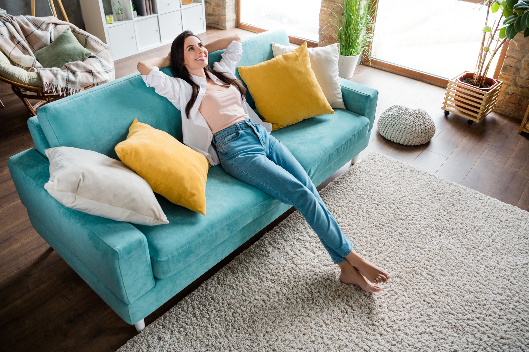 A woman is lying on a turquoise couch with her arms stretched behind her head, smiling. The living room features large windows, a light-colored rug, and several pillows in shades of white and yellow. Wooden flooring and indoor plants complement the cozy atmosphere.