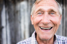 A close-up of an elderly man with short, grey hair and blue eyes, smiling warmly. He is wearing a plaid shirt and stands in front of a rustic wooden background. His face is expressive, with deep smile lines, conveying happiness and friendliness.