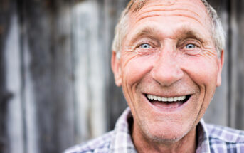 A close-up of an elderly man with short, grey hair and blue eyes, smiling warmly. He is wearing a plaid shirt and stands in front of a rustic wooden background. His face is expressive, with deep smile lines, conveying happiness and friendliness.