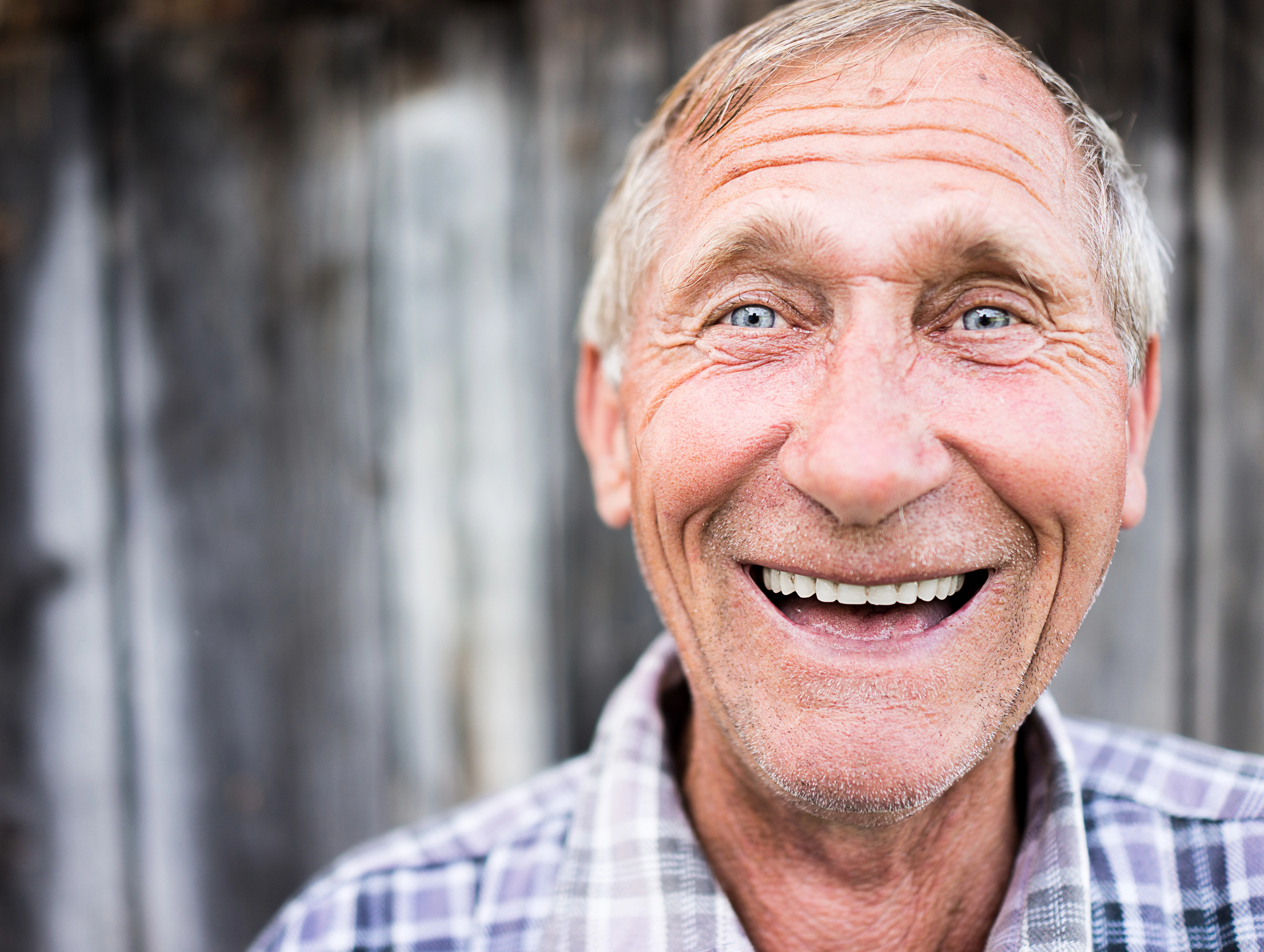 A close-up of an elderly man with short, grey hair and blue eyes, smiling warmly. He is wearing a plaid shirt and stands in front of a rustic wooden background. His face is expressive, with deep smile lines, conveying happiness and friendliness.