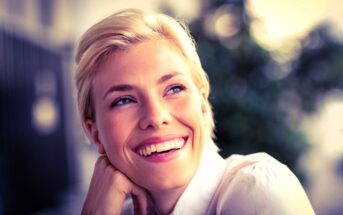 A woman with short blonde hair and blue eyes smiles brightly, looking off to the side. She rests her chin on her hand. The background is softly blurred, suggesting an outdoor setting with some greenery. She is wearing a light-colored shirt.