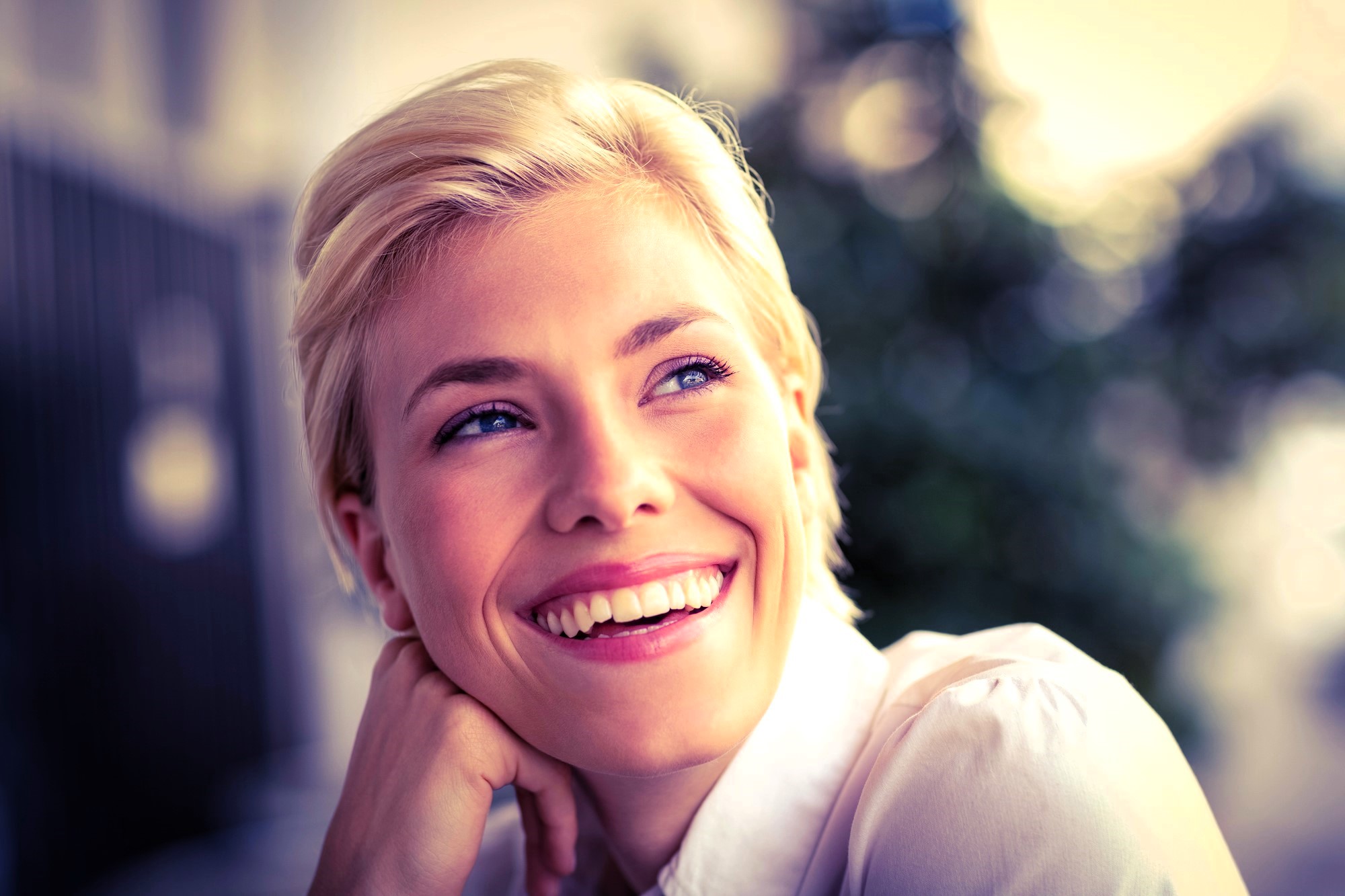 A woman with short blonde hair and blue eyes smiles brightly, looking off to the side. She rests her chin on her hand. The background is softly blurred, suggesting an outdoor setting with some greenery. She is wearing a light-colored shirt.