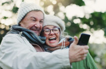 An elderly couple in warm clothing takes a cheerful selfie outdoors. Both are wearing winter hats and scarves, with greenery visible in the background. They are smiling and appear joyful.