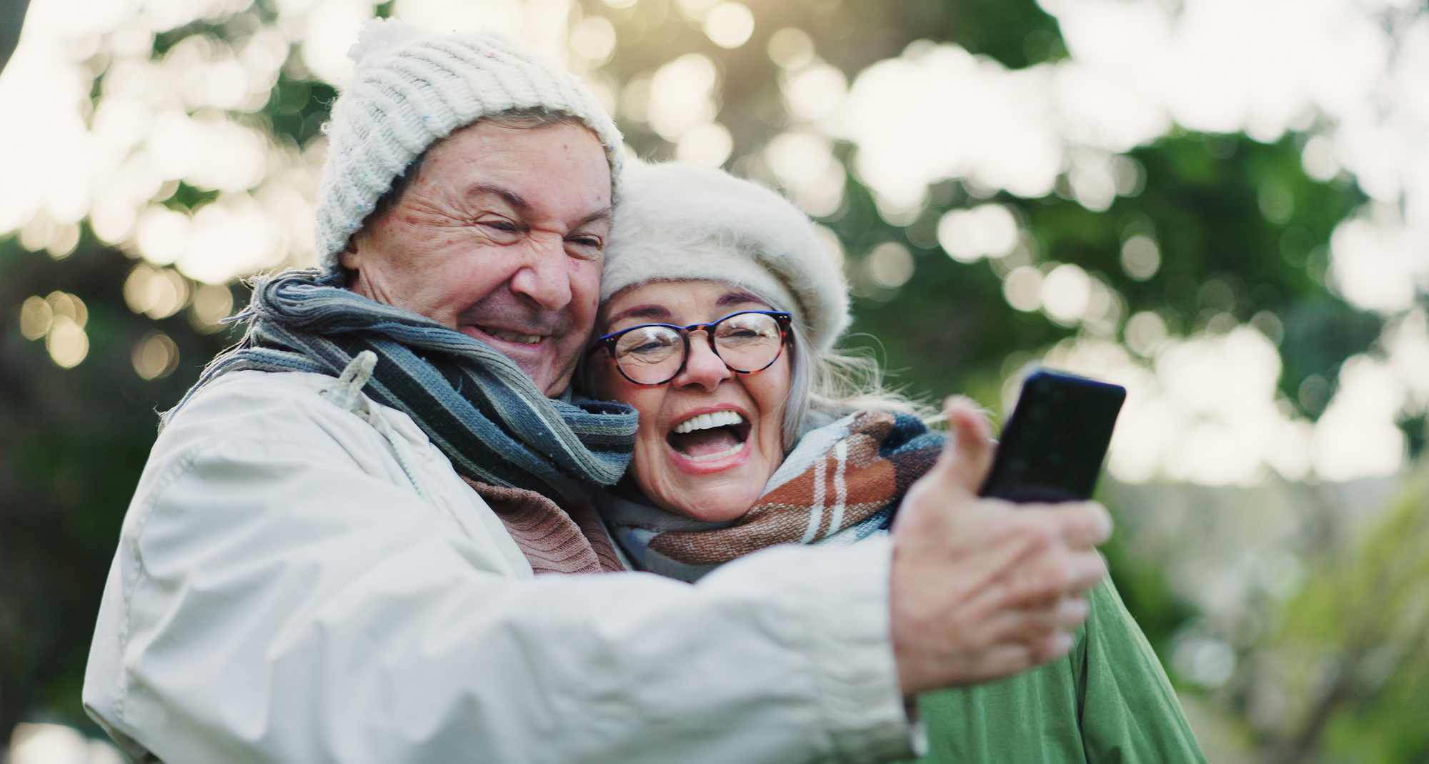 An elderly couple in warm clothing takes a cheerful selfie outdoors. Both are wearing winter hats and scarves, with greenery visible in the background. They are smiling and appear joyful.