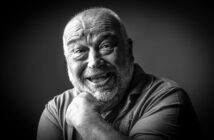 A black-and-white portrait of an elderly man with a bald head and beard smiling broadly. He supports his chin with one hand while looking directly at the camera. The background is dark, highlighting his expressive and joyful facial expression.