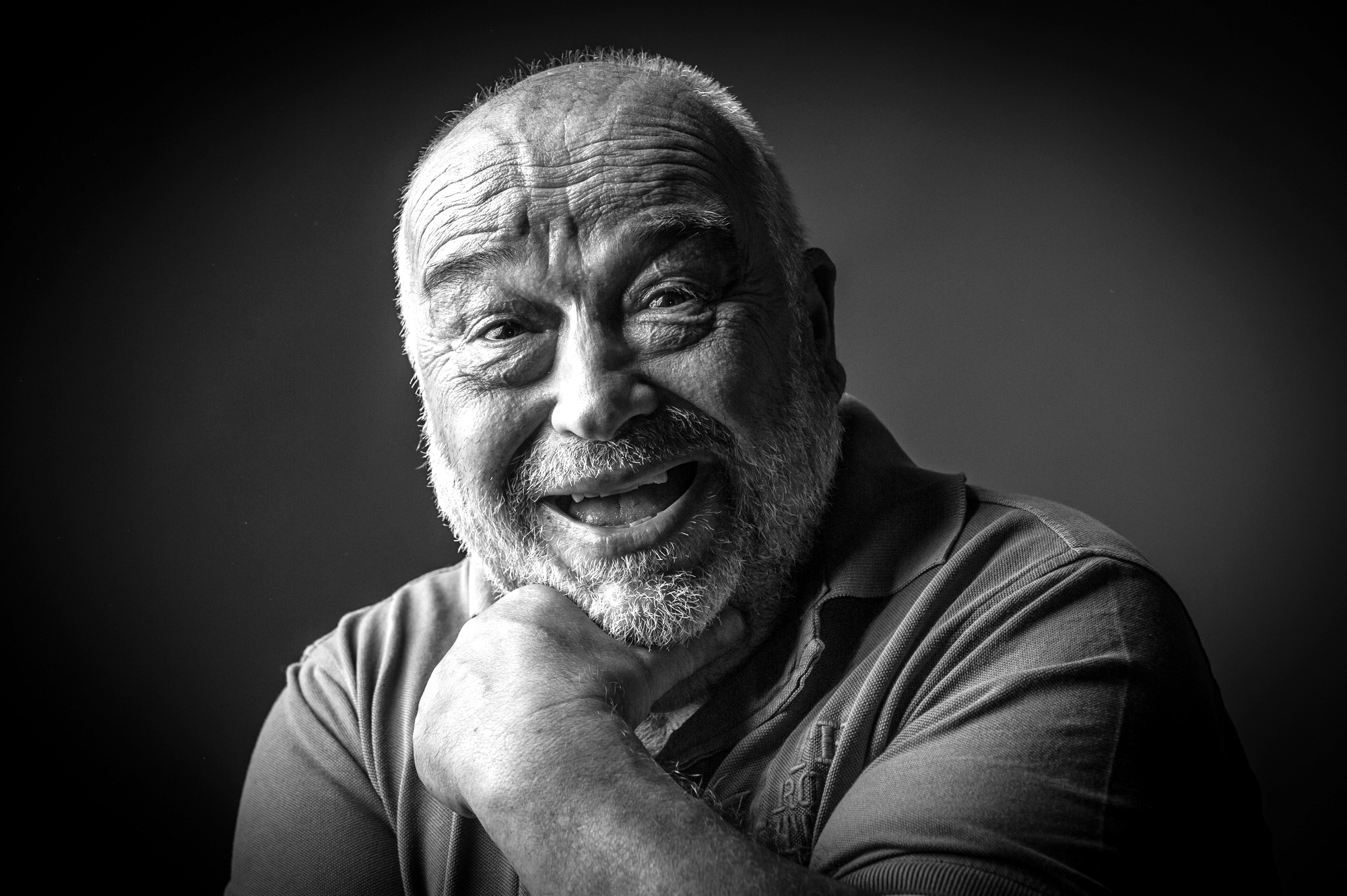 A black-and-white portrait of an elderly man with a bald head and beard smiling broadly. He supports his chin with one hand while looking directly at the camera. The background is dark, highlighting his expressive and joyful facial expression.