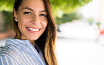 A woman with long brown hair wearing a light blue and white striped shirt is smiling at the camera. She is standing outdoors with blurred greenery in the background. The lighting is bright, highlighting her cheerful expression.