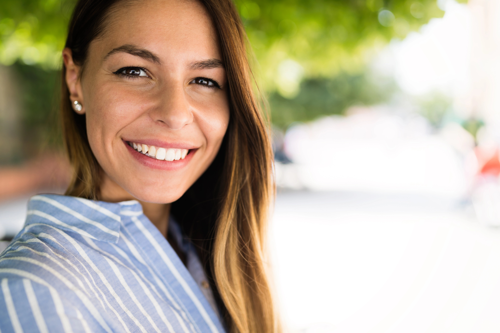 A woman with long brown hair wearing a light blue and white striped shirt is smiling at the camera. She is standing outdoors with blurred greenery in the background. The lighting is bright, highlighting her cheerful expression.