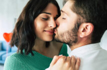 Close-up of a couple sharing an intimate moment. The man, with short hair and a beard, gently kisses the cheek of the smiling woman, who has shoulder-length dark hair and wears a green top. They appear to be in a cozy indoor setting.