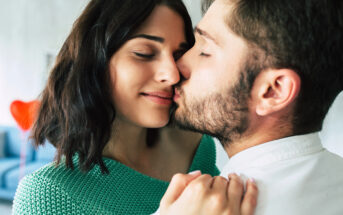 Close-up of a couple sharing an intimate moment. The man, with short hair and a beard, gently kisses the cheek of the smiling woman, who has shoulder-length dark hair and wears a green top. They appear to be in a cozy indoor setting.