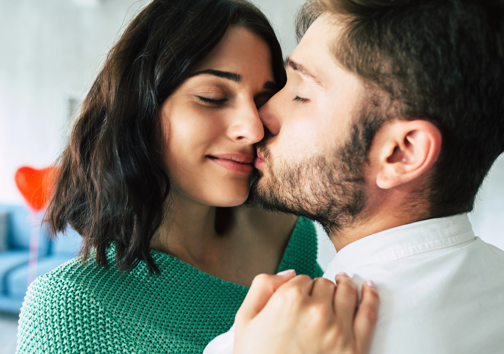 Close-up of a couple sharing an intimate moment. The man, with short hair and a beard, gently kisses the cheek of the smiling woman, who has shoulder-length dark hair and wears a green top. They appear to be in a cozy indoor setting.