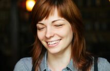 A young woman with light brown hair and bangs is smiling with her eyes closed. She is wearing a checkered shirt and is in a dimly lit setting, possibly a café or a similar cozy environment. The background is blurred, emphasizing her content expression.