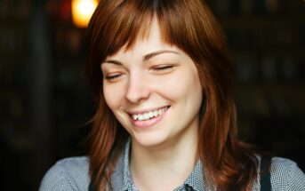 A young woman with light brown hair and bangs is smiling with her eyes closed. She is wearing a checkered shirt and is in a dimly lit setting, possibly a café or a similar cozy environment. The background is blurred, emphasizing her content expression.