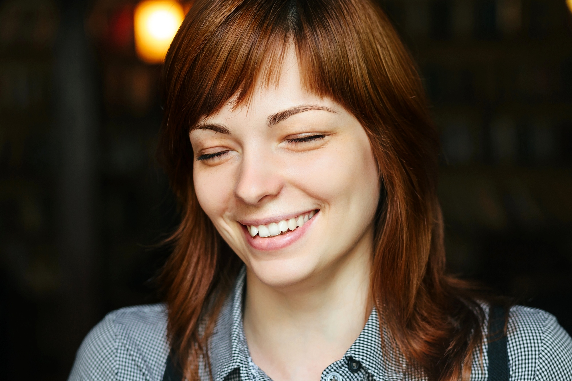 A young woman with light brown hair and bangs is smiling with her eyes closed. She is wearing a checkered shirt and is in a dimly lit setting, possibly a café or a similar cozy environment. The background is blurred, emphasizing her content expression.