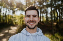 A man with dark hair and a beard smiles at the camera while standing outdoors. He is wearing a light grey hoodie and the background features sunlight filtering through tall trees. The setting appears to be a forest or wooded area.