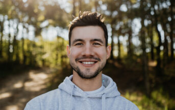 A man with dark hair and a beard smiles at the camera while standing outdoors. He is wearing a light grey hoodie and the background features sunlight filtering through tall trees. The setting appears to be a forest or wooded area.