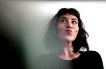 A woman with shoulder-length dark hair and wearing a black top looks up and to the side with a thoughtful expression, pursing her lips. The background is a plain, light-colored wall.