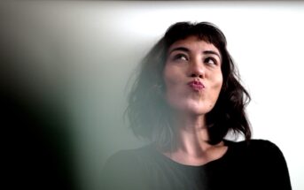 A woman with shoulder-length dark hair and wearing a black top looks up and to the side with a thoughtful expression, pursing her lips. The background is a plain, light-colored wall.