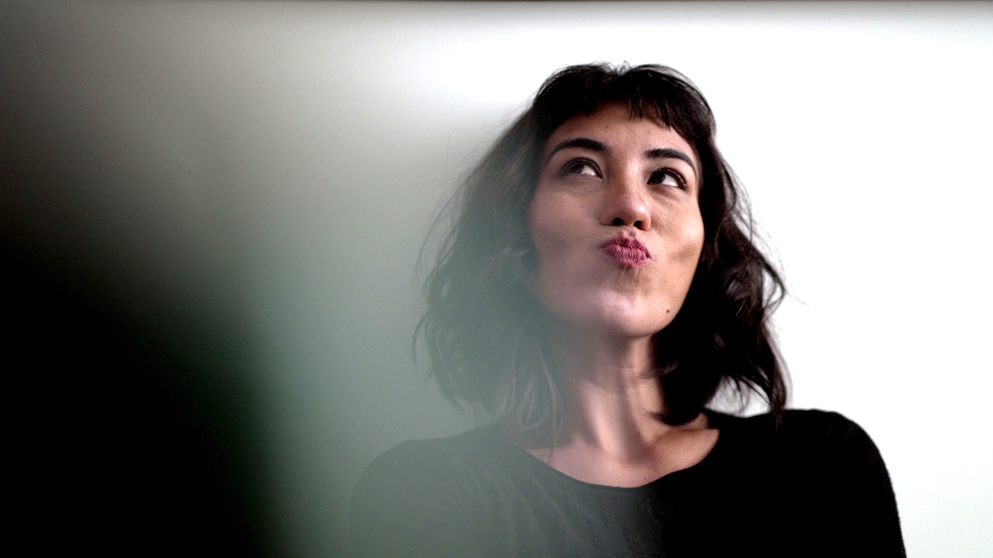 A woman with shoulder-length dark hair and wearing a black top looks up and to the side with a thoughtful expression, pursing her lips. The background is a plain, light-colored wall.