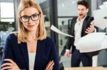 A woman with blonde hair and glasses confidently stands with her arms crossed and a slight smile in an office setting. In the background, a man in a suit is angrily throwing papers in the air. The scene looks tense but she appears composed.