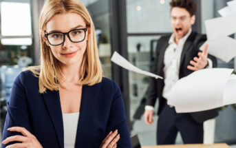 A woman with blonde hair and glasses confidently stands with her arms crossed and a slight smile in an office setting. In the background, a man in a suit is angrily throwing papers in the air. The scene looks tense but she appears composed.