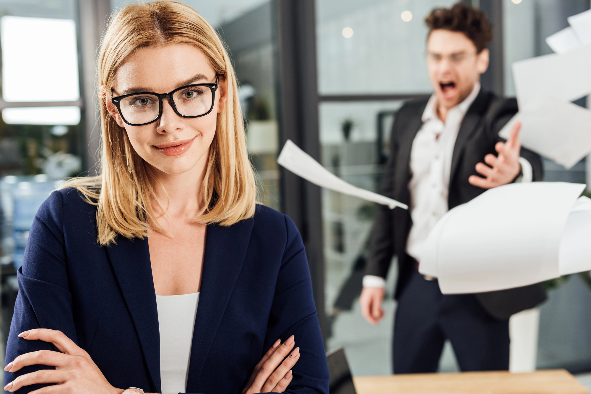 A woman with blonde hair and glasses confidently stands with her arms crossed and a slight smile in an office setting. In the background, a man in a suit is angrily throwing papers in the air. The scene looks tense but she appears composed.