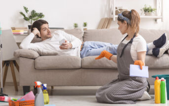 A woman kneels on the floor with cleaning supplies and looks at a man lying on a couch using his phone. She wears an apron and rubber gloves, holding a cloth. The man seems relaxed while the woman is engaged in cleaning tasks. Various cleaning products are nearby.