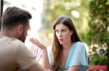 A woman with long brown hair wearing a light blue top is talking to a man with short hair and a beard, who is facing away from the camera. They are sitting outdoors, and the background is a blurred view of greenery and buildings.