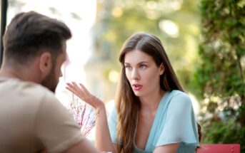 A woman with long brown hair wearing a light blue top is talking to a man with short hair and a beard, who is facing away from the camera. They are sitting outdoors, and the background is a blurred view of greenery and buildings.