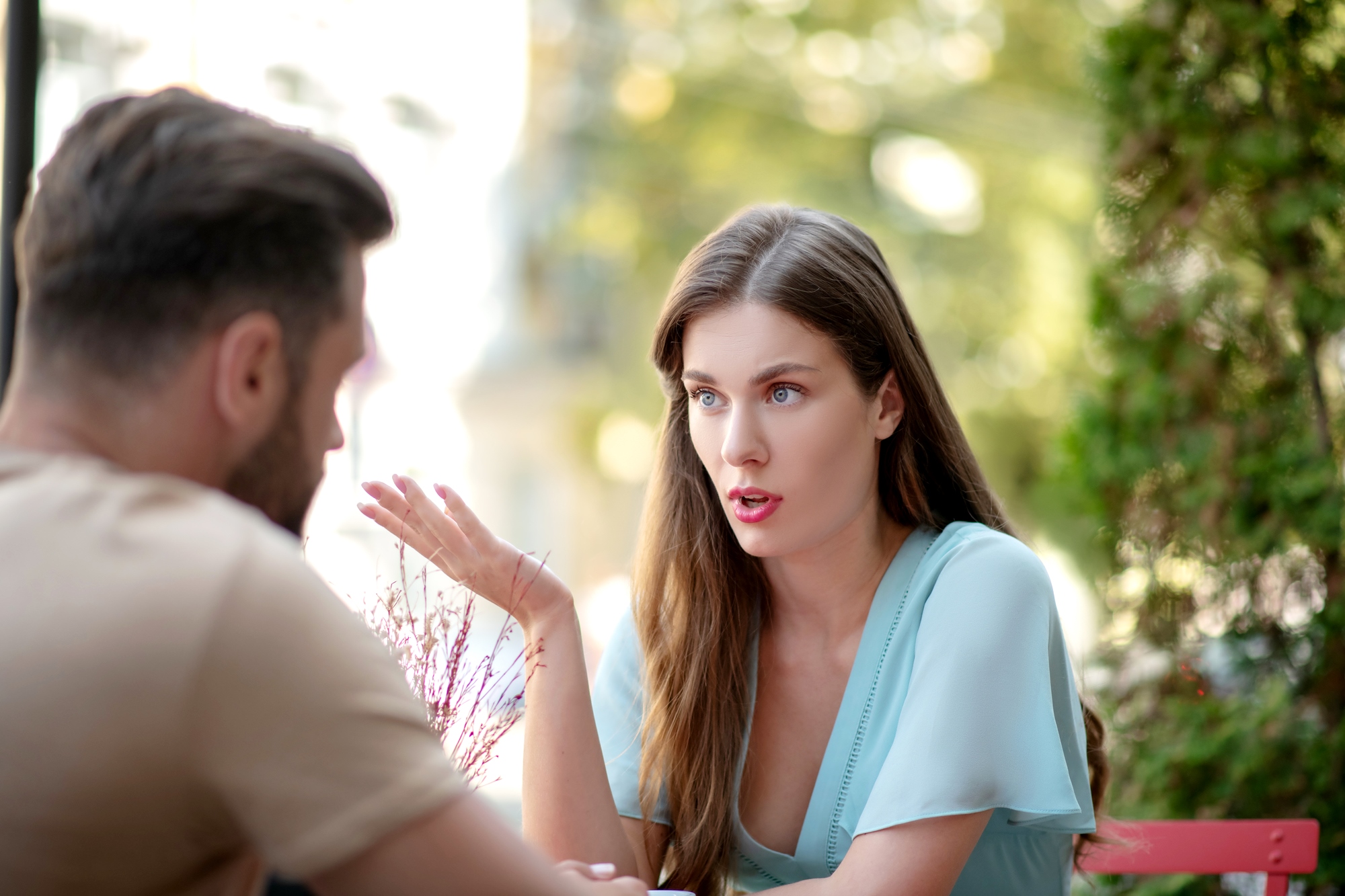 A woman with long brown hair wearing a light blue top is talking to a man with short hair and a beard, who is facing away from the camera. They are sitting outdoors, and the background is a blurred view of greenery and buildings.