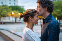 A man gently kisses a woman's forehead as they stand close, facing each other, outdoors in an urban setting. The woman smiles, eyes closed, wearing a white top, while the man wears a blue shirt with rolled-up sleeves. The background is softly lit with evening lights.