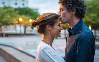 A man gently kisses a woman's forehead as they stand close, facing each other, outdoors in an urban setting. The woman smiles, eyes closed, wearing a white top, while the man wears a blue shirt with rolled-up sleeves. The background is softly lit with evening lights.