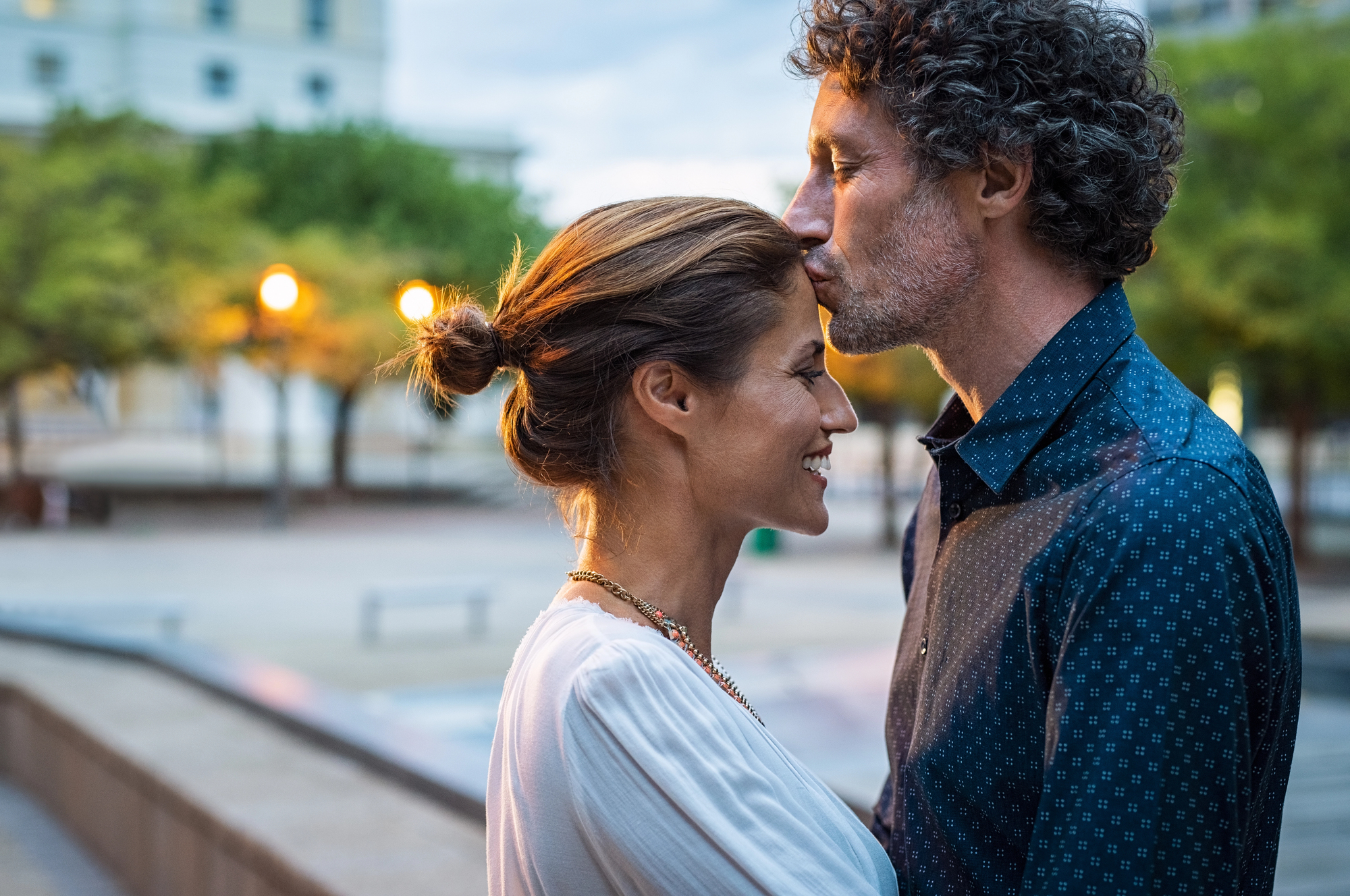 A man gently kisses a woman's forehead as they stand close, facing each other, outdoors in an urban setting. The woman smiles, eyes closed, wearing a white top, while the man wears a blue shirt with rolled-up sleeves. The background is softly lit with evening lights.