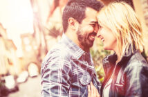 A happy couple stands close together in a sunlit street. The man, with a beard and plaid shirt, smiles with his eyes closed, touching foreheads with the woman, who has blonde hair and is wearing a leather jacket. Both appear joyful and affectionate.