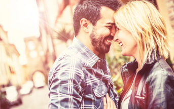 A happy couple stands close together in a sunlit street. The man, with a beard and plaid shirt, smiles with his eyes closed, touching foreheads with the woman, who has blonde hair and is wearing a leather jacket. Both appear joyful and affectionate.