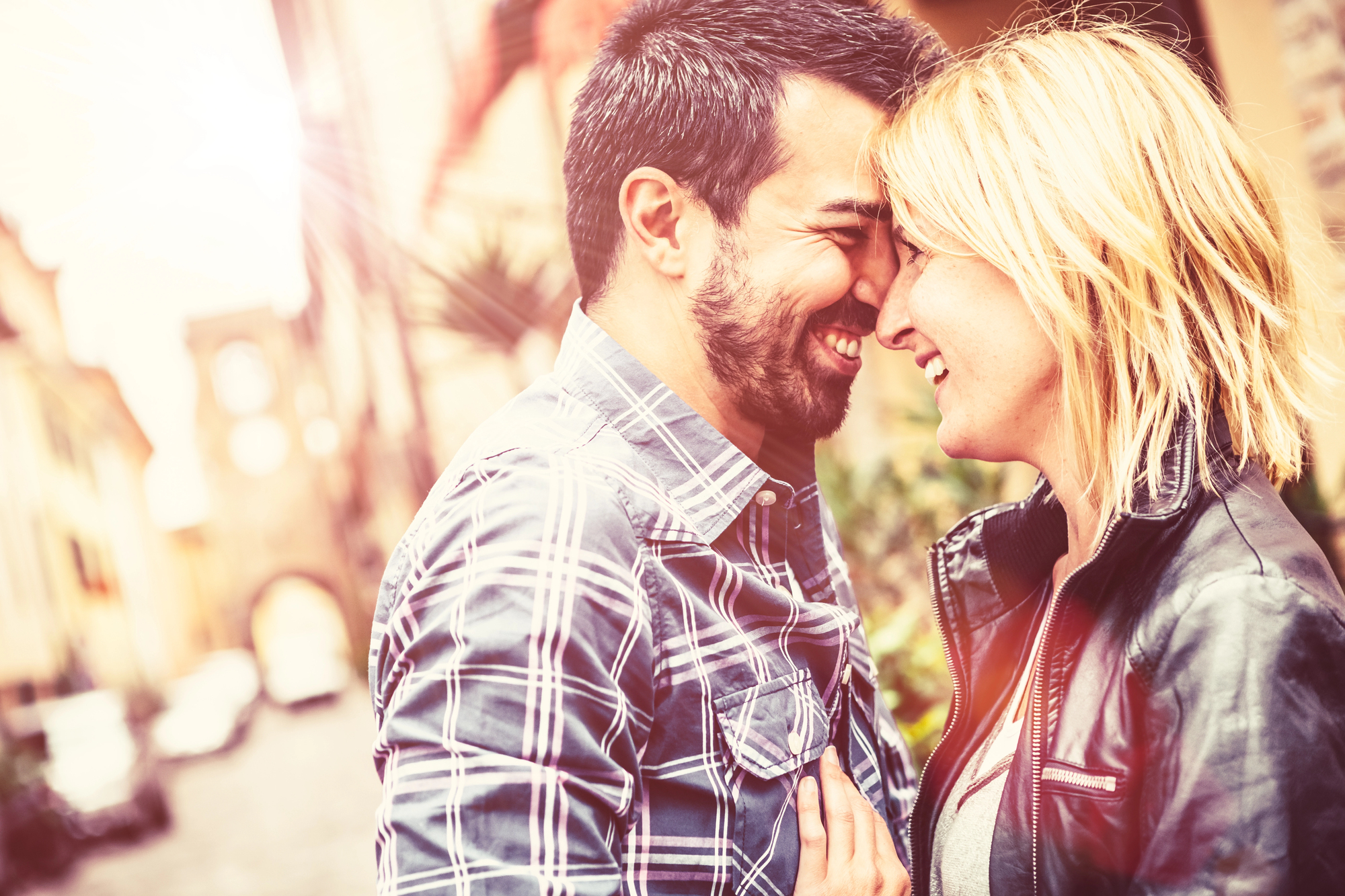 A happy couple stands close together in a sunlit street. The man, with a beard and plaid shirt, smiles with his eyes closed, touching foreheads with the woman, who has blonde hair and is wearing a leather jacket. Both appear joyful and affectionate.