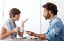 A woman and a man sit across from each other at a table, engaging in a conversation. The woman, holding a coffee cup, gestures with her hand, while the man listens attentively with an open posture. Both appear to be in a casual setting, possibly a café.