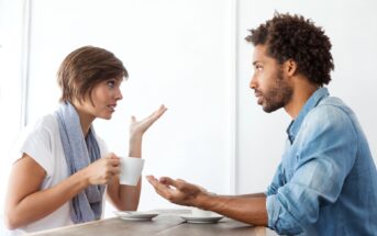 A woman and a man sit across from each other at a table, engaging in a conversation. The woman, holding a coffee cup, gestures with her hand, while the man listens attentively with an open posture. Both appear to be in a casual setting, possibly a café.