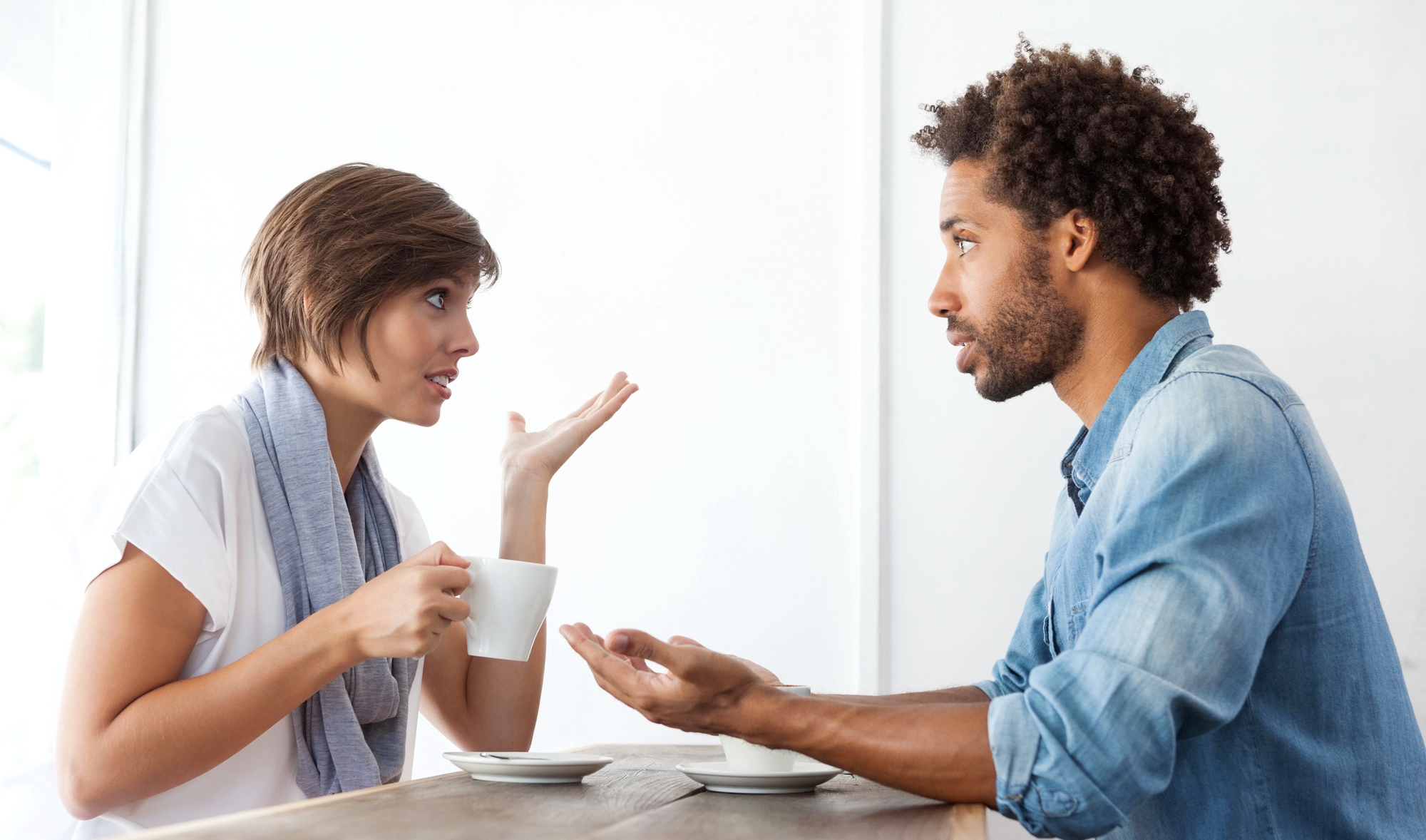 A woman and a man sit across from each other at a table, engaging in a conversation. The woman, holding a coffee cup, gestures with her hand, while the man listens attentively with an open posture. Both appear to be in a casual setting, possibly a café.