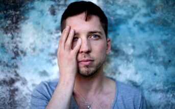 A man with short dark hair and a light stubble stands against a textured blue and gray background. He wears a light gray shirt and a small cross necklace, and he covers his right eye with his right hand, looking directly at the camera with his left eye visible.