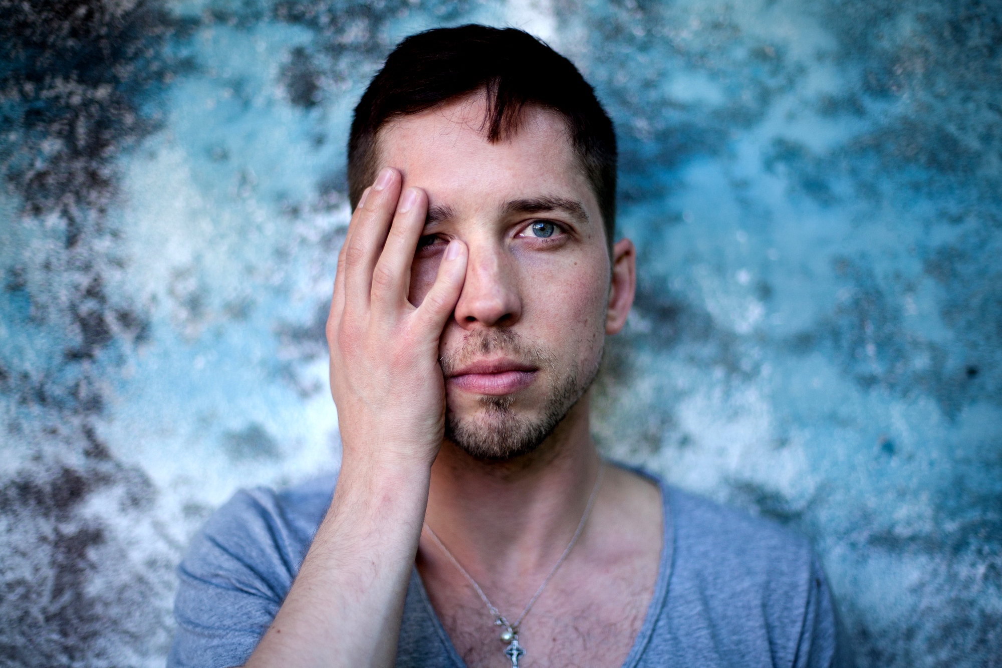 A man with short dark hair and a light stubble stands against a textured blue and gray background. He wears a light gray shirt and a small cross necklace, and he covers his right eye with his right hand, looking directly at the camera with his left eye visible.