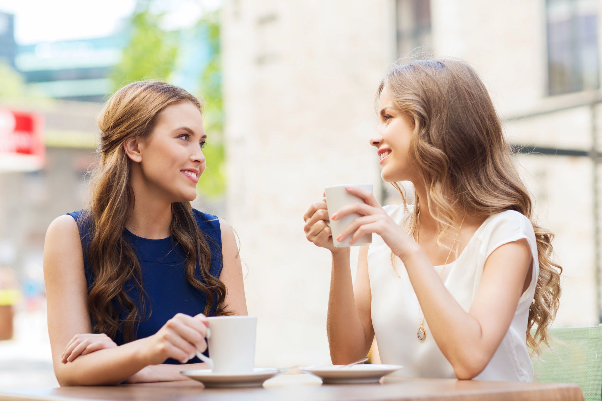 Two women with long hair sit at an outdoor cafe table, smiling and holding white mugs. One wears a navy blue top, and the other has a white top. There are blurred city buildings in the background with a sunlit ambiance.