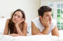 A woman and a man lie on a bed, propping their heads on their hands and looking away in opposite directions with contemplative expressions. They appear to be in a bright, airy room with white bedding and a large window in the background.