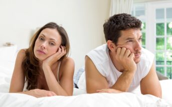 A woman and a man lie on a bed, propping their heads on their hands and looking away in opposite directions with contemplative expressions. They appear to be in a bright, airy room with white bedding and a large window in the background.