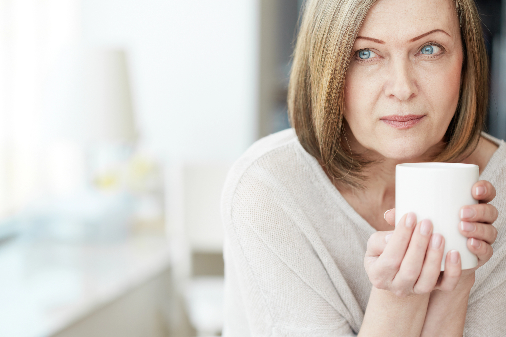 A woman with short, light brown hair is holding a white mug with both hands and looking thoughtfully into the distance. She is wearing a light-colored, long-sleeved top. The background is softly blurred, creating a calm and serene atmosphere.