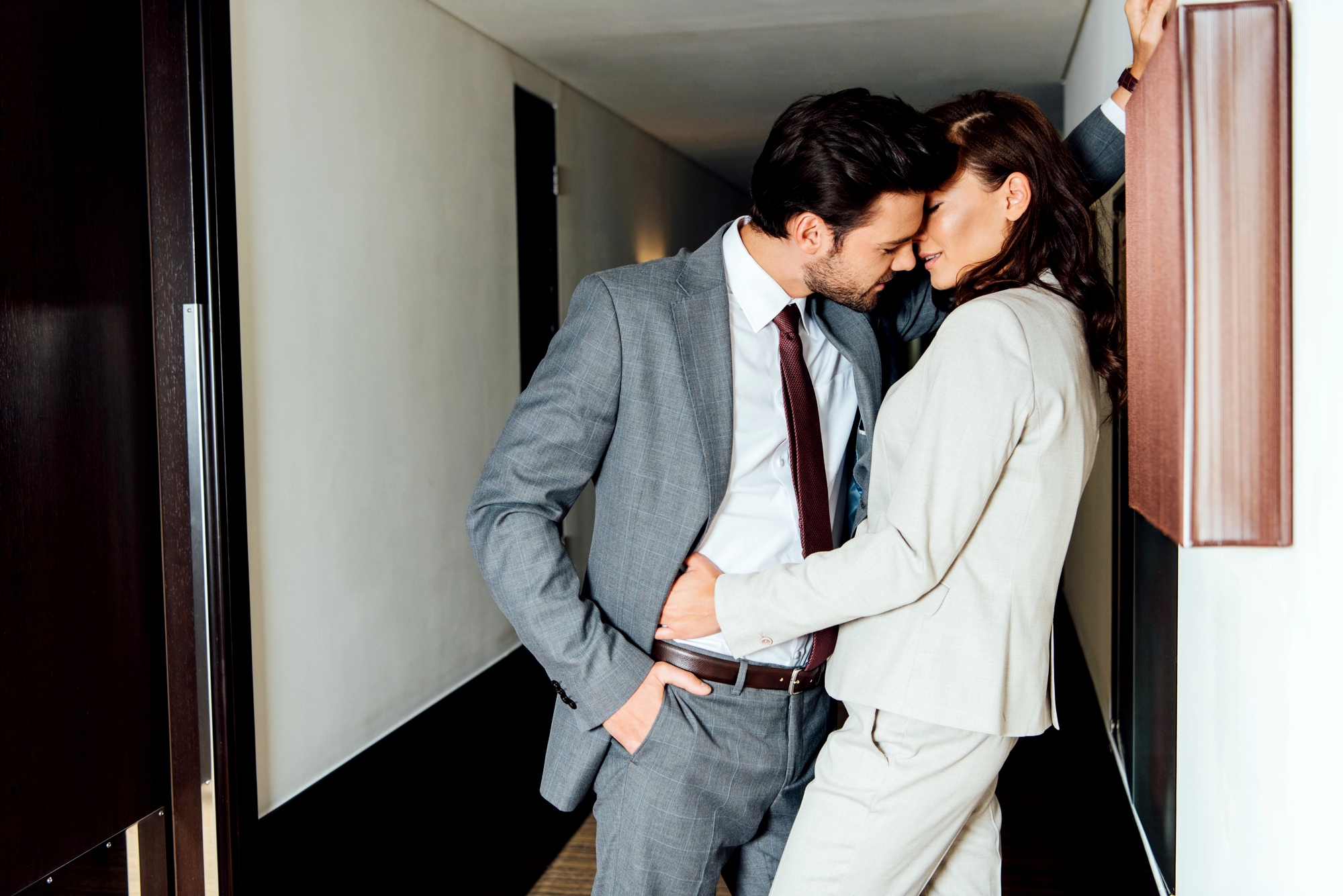 A man in a gray suit and a woman in a light-colored suit are standing close to each other in a hallway, with their foreheads touching affectionately. The man has his hand in his pocket while the woman leans against the wall. Both seem to be sharing an intimate moment.